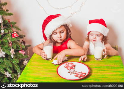 Two small Santas are drinking milk and eating colorful gingerbreads. Two girls near Christmas tree