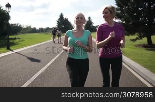 Two smiling athletic senior women runnining in park on a sunny day. Beautiful adult blonde female runners jogging during outdoor workout in park. Running fit females in sport clothes enjoying training outdoors. Slow motion. Steadicam stabilized shot.