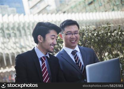 Two smiling businessmen looking at laptop together outdoors in Beijing