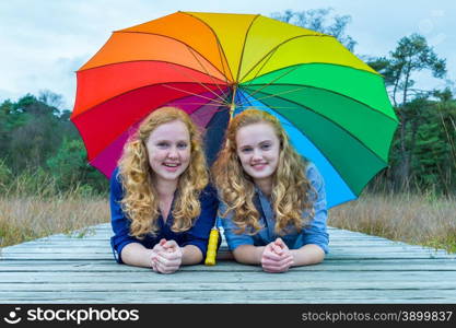 Two smiling caucasian teenage girlfriends lying in nature under colorful umbrella
