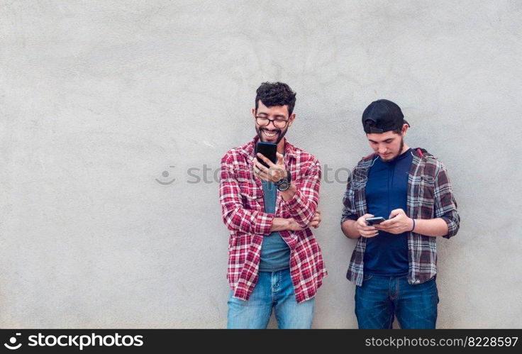 Two smiling friends leaning on a wall checking their cell phones, Friends leaning on a wall texting on their phones. Friend showing cell phone to his friend, Smiling friends checking cell phones