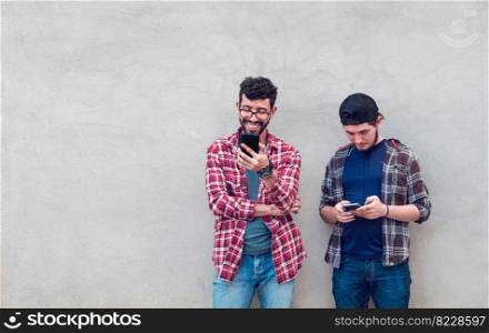 Two smiling friends leaning on a wall checking their cell phones, Friends leaning on a wall texting on their phones. Friend showing cell phone to his friend, Smiling friends checking cell phones