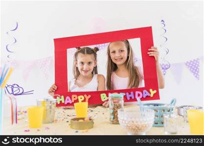 two smiling girl holding happy birthday frame with different foods table