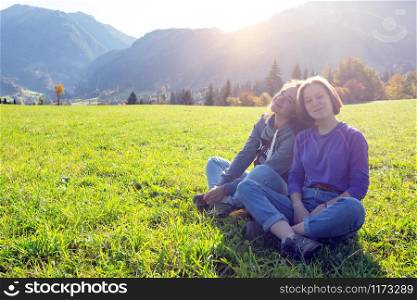 Two smiling girls are sitting on a green meadow and mountains in the background