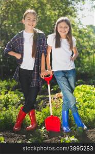 Two smiling girls in rubber boots posing at garden with shovel