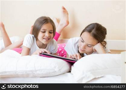 Two smiling girls lying on bed and browsing internet on digital tablet