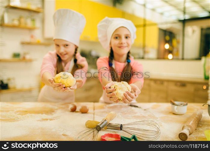 Two smiling little girls cooks in caps shows dough balls, cookies preparation on the kitchen. Kids cooking pastry, children chefs preparing cake