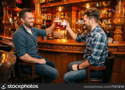 Two smiling male friends drinks beer at the counter in pub. Bearded men with mugs having fun in bar, good friends. Two male friends drinks beer at the counter in pub