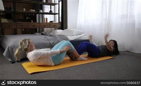 Two smiling middle-aged sisters doing abdominal crunches press exercise on the mat in domestic interior. Sporty attractive women working out together at home and doing sit-up exercise. Slo mo. Dolly
