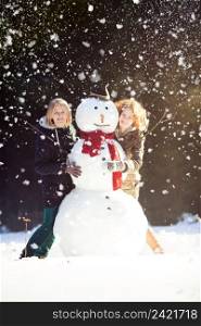 Two smiling pretty girls posing with a snowman in forest, it’s snowing