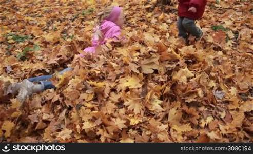 Two smiling siblings lying on their bellies in a pile of fallen autumn leaves, throwing them up in the air in public park. Cute toddler boy and his teenage sister having fun and playing with yellow and orange foliage outdoors in autumn time.