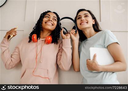Two smiling women in headphones listening to music indoors. Pretty girlfriends in earphones relax in the room, music lovers resting. Smiling women in headphones listening to music