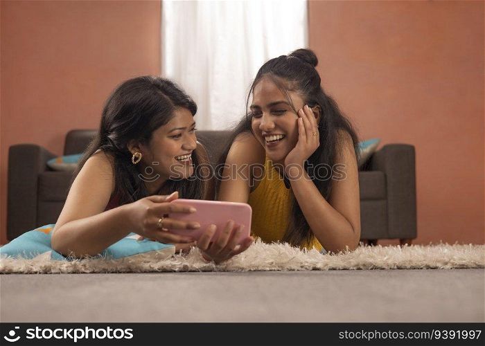 Two smiling women together watching video on mobile phone while lying on floor in living room