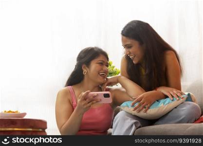 Two smiling women together watching video on mobile phone while sitting in living room