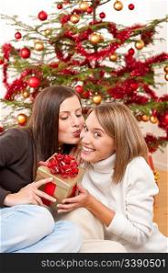 Two smiling women with Christmas present kissing in front of tree