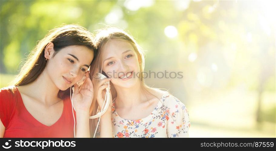 Two Smiling Young Girls with Pleasure Listening to Music on Headphones on a fine Summer Day