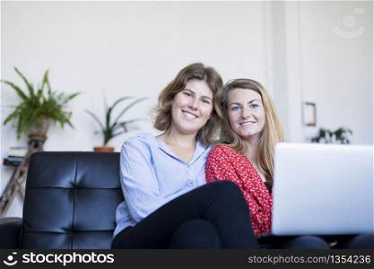 Two smiling young woman sitting on the sofa with laptop.