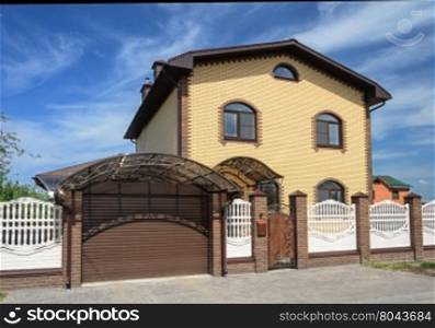 Two-storeyed yellow brick cottage with white concrete fence and metal gate, sunny summer day