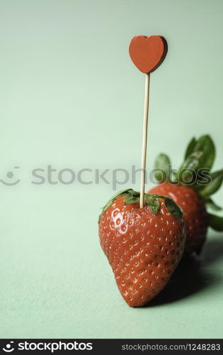 Two strawberries and a heart-shaped toothpick stuck in it on a green seamless background. Ripe strawberries. Sweet summer fruits. Valentine day card.