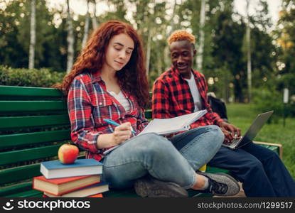 Two students drinks coffee on the bench in summer park. Male and female white teenagers relax outdoors. Two students drinks coffee on the bench in park