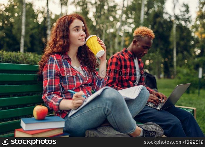 Two students drinks coffee on the bench in summer park. Male and female white teenagers relax outdoors. Two students drinks coffee on the bench in park