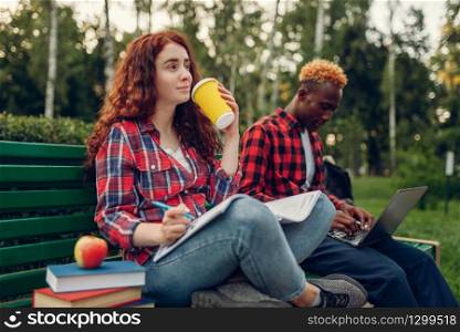 Two students drinks coffee on the bench in summer park. Male and female white teenagers relax outdoors. Two students drinks coffee on the bench in park