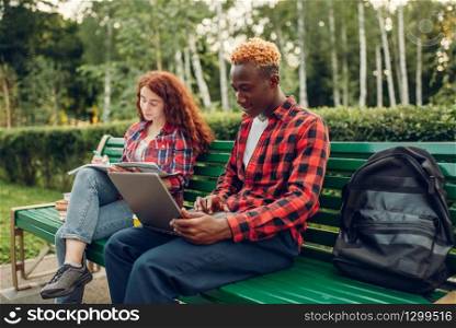 Two students studying on the bench in summer park. Male and female white teenagers relax outdoors. Two students studying on the bench in summer park