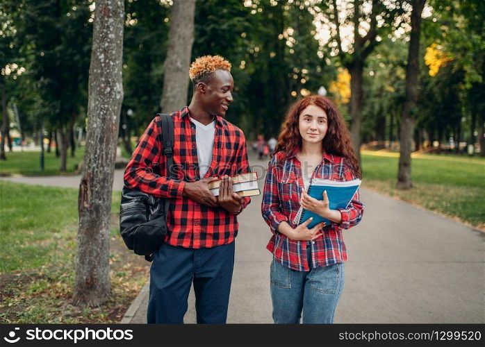 Two students walk on the sidewalk in summer park. Male and female white teenagers relax outdoors. Two students walk on the sidewalk in summer park