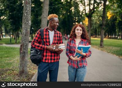 Two students walk on the sidewalk in summer park. Male and female white teenagers relax outdoors. Two students walk on the sidewalk in summer park