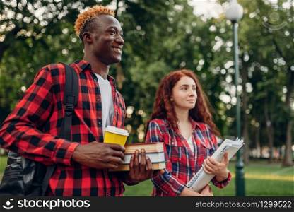 Two students walk on the sidewalk in summer park. Male and female white teenagers outdoors. Two students walk on the sidewalk in summer park