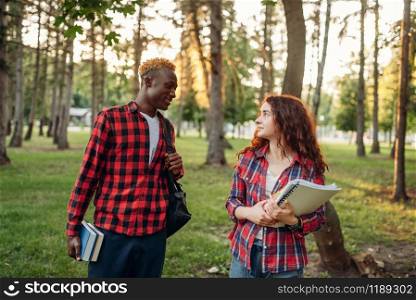 Two students walking on lawn in summer park. Male and female white teenagers studying outdoors. Two students walking on lawn in summer park