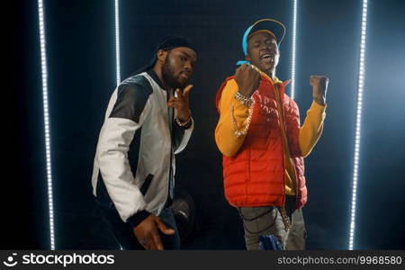 Two stylish rappers poses in glowing cube, studio with dark background. Hip-hop performers, break-dancers. Two stylish rappers poses in glowing cube