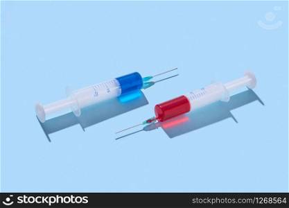 Two surgical disposable syringes 20 ml with red and blue serum or vaccine for an intravenous injection on a pastel blue background with hard shadow, copy space.. Plastic syringes of red and blue vaccine with shadows.