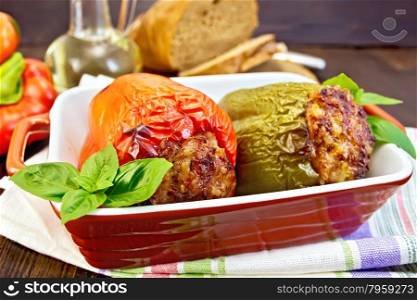 Two sweet peppers stuffed with meat and rice with basil leaves in a brown roasting pan on a napkin on a wooden boards background