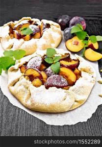Two sweet pies with plum, sugar and cardamom on parchment, sprigs of green mint on a dark wooden board background