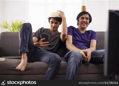 Two teenage boys having fun while sitting on sofa with bowl and plastic glass on head
