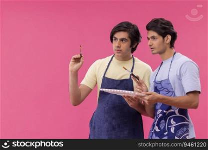 Two teenage boys holding paintbrush and colour palette against pink background