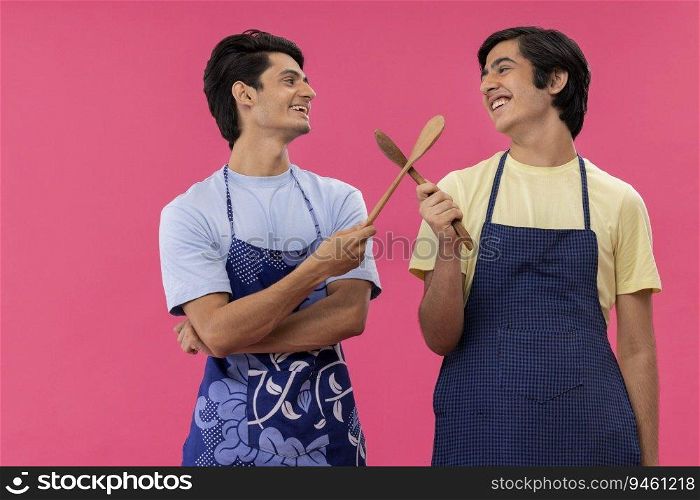Two teenage boys in chef apron standing with spatulas against pink background