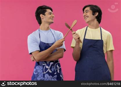 Two teenage boys in chef apron standing with spatulas against pink background