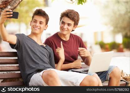 Two Teenage Boys Sitting On Bench Taking Selfie In Park