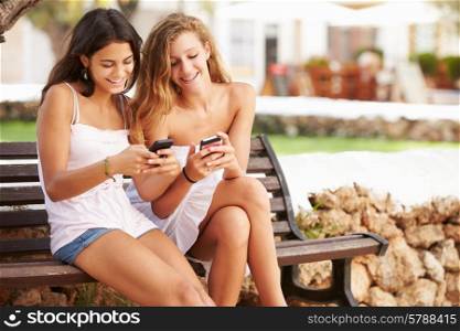Two Teenage Girl Using Mobile Phone Sitting On Park Bench