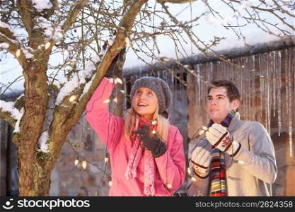 Two Teenage Girls Hanging Fairy Lights In Tree With Icicles In Foreground