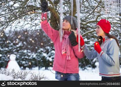 Two Teenage Girls Hanging Fairy Lights In Tree With Icicles In Foreground