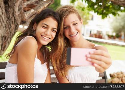 Two Teenage Girls Sitting On Bench Taking Selfie In Park