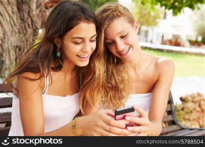 Two Teenage Girls Using Mobile Phone Sitting On Park Bench