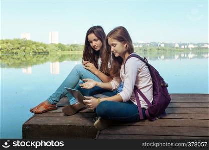 Two teenager smiling girls with backpacks sitting on a pier at the river bank and the city in the background
