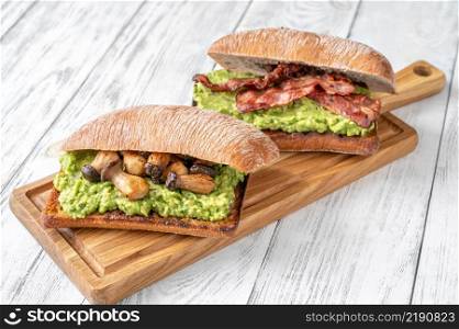 Two toasts with guacamole, mushrooms and bacon on cutting board