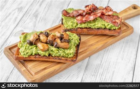 Two toasts with guacamole, mushrooms and bacon on cutting board