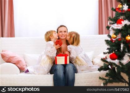 Two twins daughters presenting gift to mother and kissing her near Christmas tree&#xA;