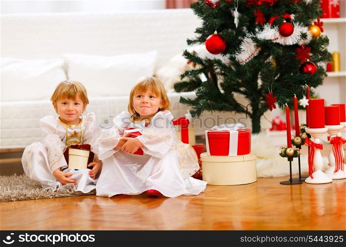 Two twins girl sitting with presents near Christmas tree